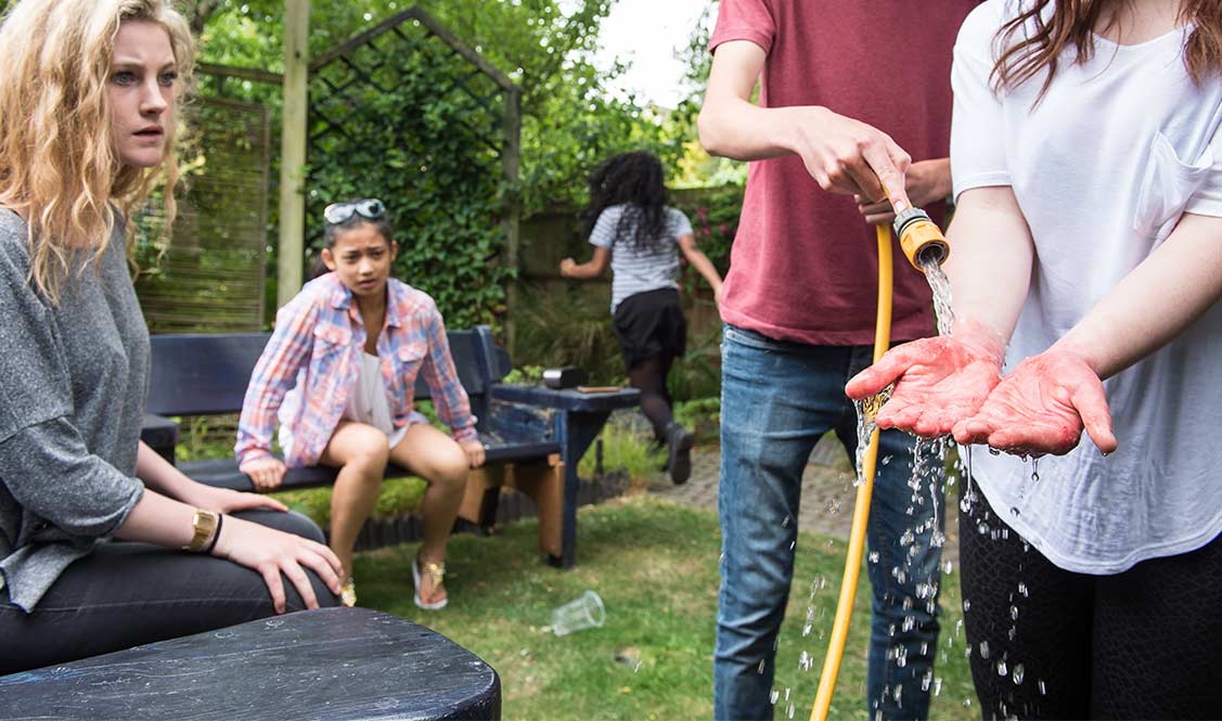 Sam's friend uses running water from a hose to cool Sam's burnt hands. Mae ffrind Sam yn defnyddio dŵr rhedeg o bibell i oeri dwylo llosg Sam.