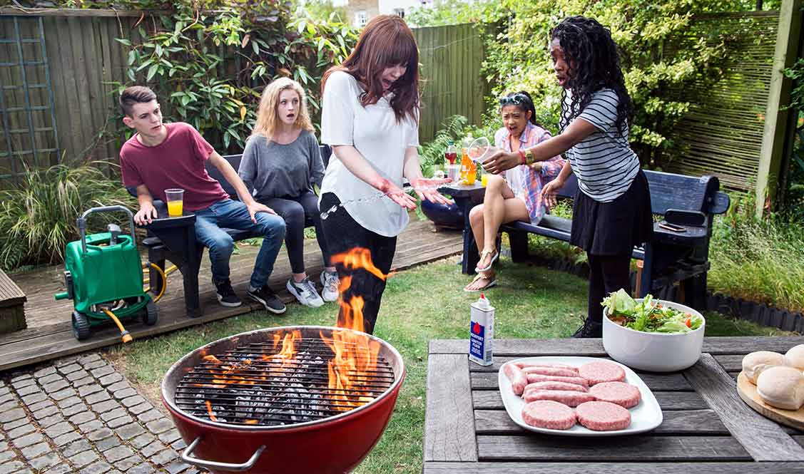 Sam stands behind a lit barbecue looking at her burnt hands, while her friends look on shocked