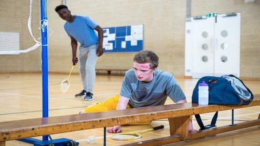 Simon is on the floor after falling and hitting his head on a bench, Nick looks concerned. Mae Simon ar y llawr ar ôl cwympo a tharo ei ben ar fainc, mae Nick yn edrych yn bryderus. 